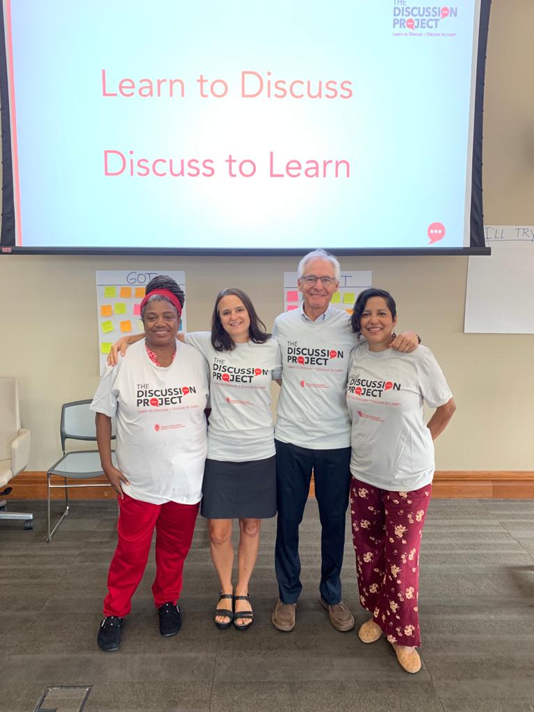 Four Discussion Project instructors smile in front of a projector screen that says "Learn to Discuss, Discuss to Learn"